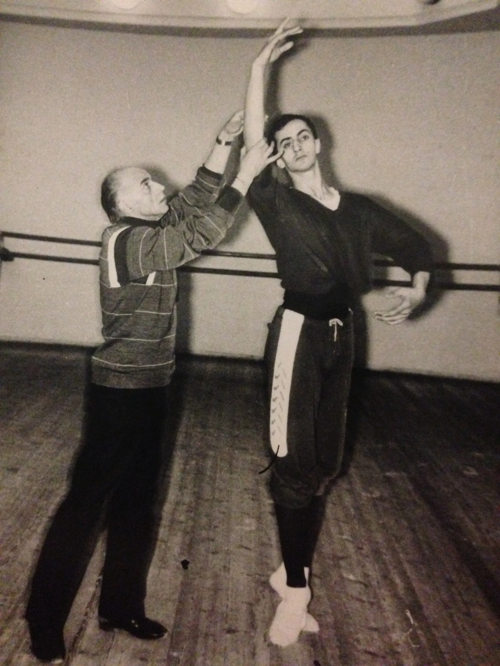À la barre, un professeur corrige le bras d'un jeune danseur, en noir et blanc.