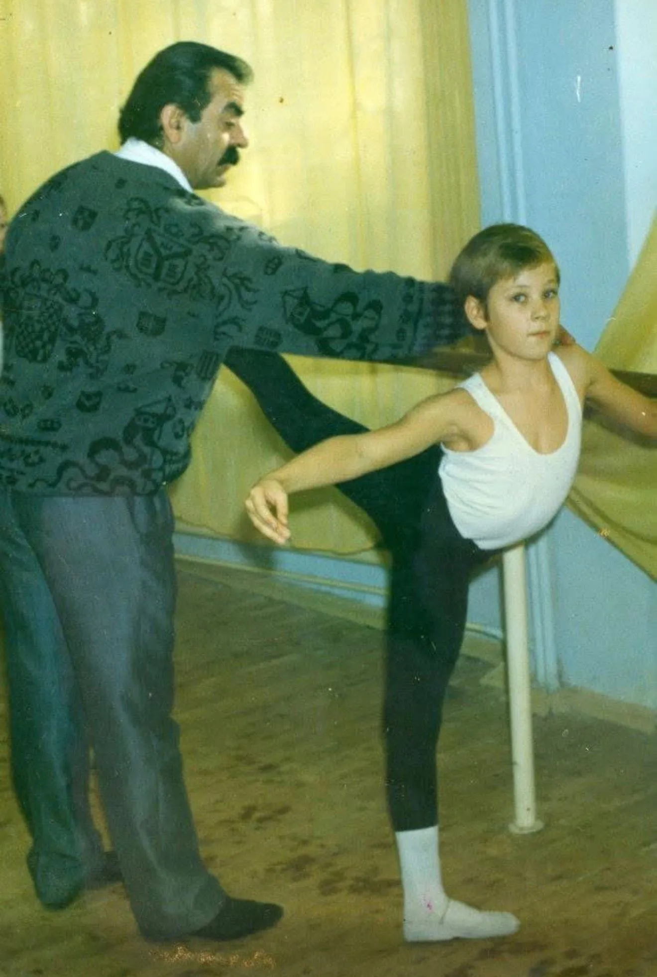 Un professeur guide la jambe d'un jeune élève à la barre, tirage couleur d'époque.