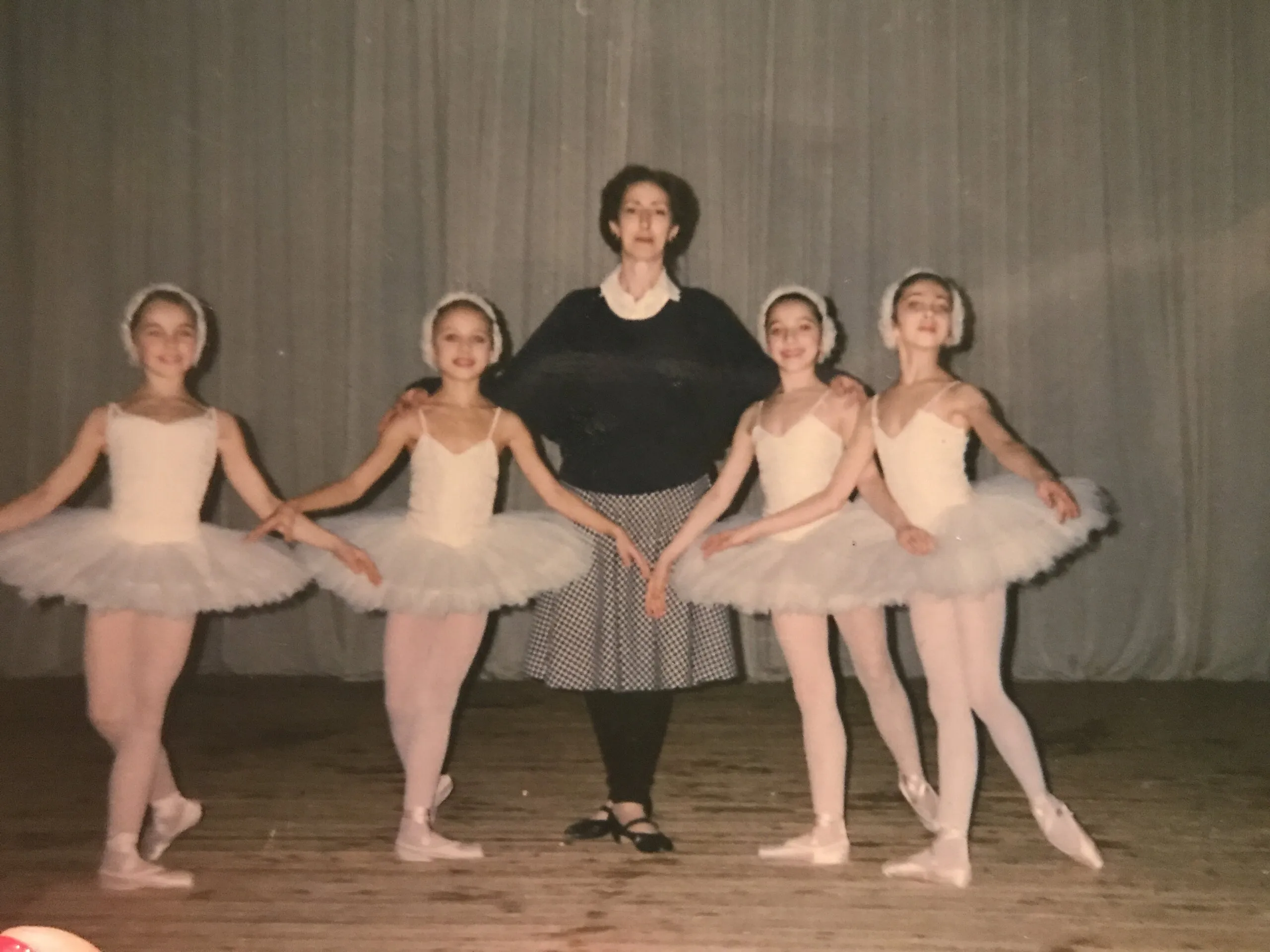 Quatre petites élèves en tutus blancs encadrent leur professeure sur une scène.
