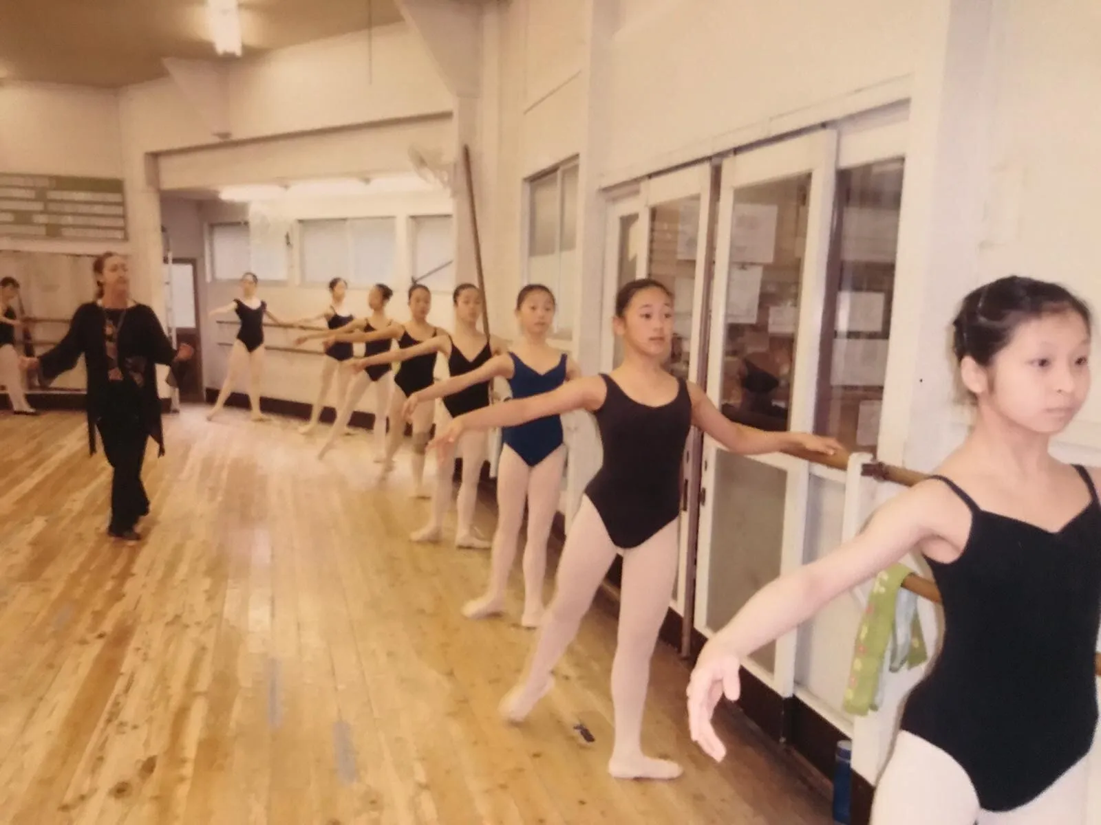 À la barre, les élèves en justaucorps sombres, la professeure passe derrière elles.