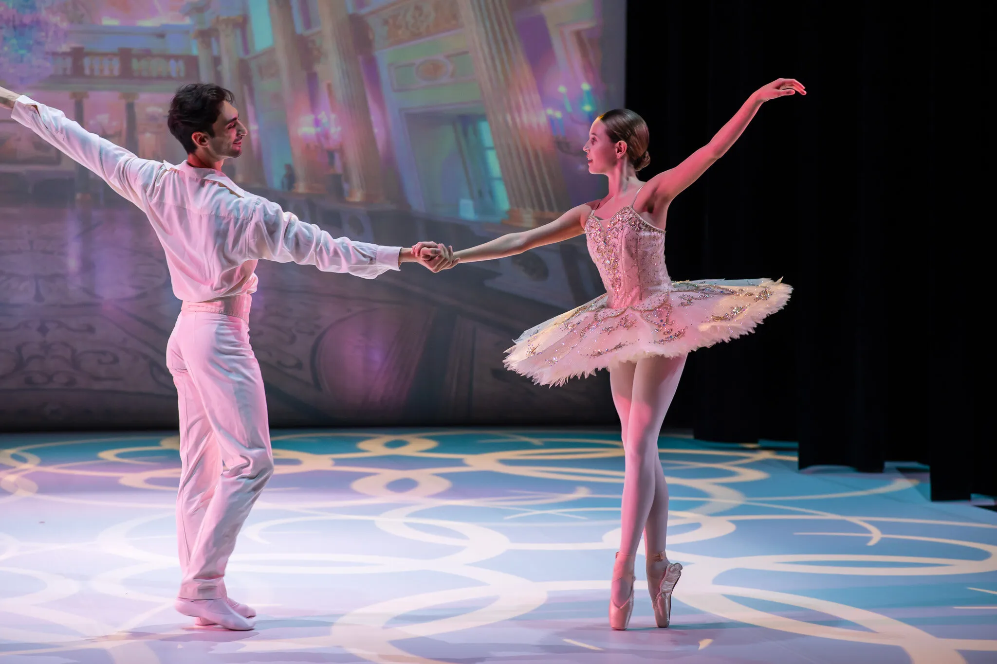 During the dress rehearsal, a young ballerina in a pale pink tutu on pointe, her hand held at arm's length by her partner in white.