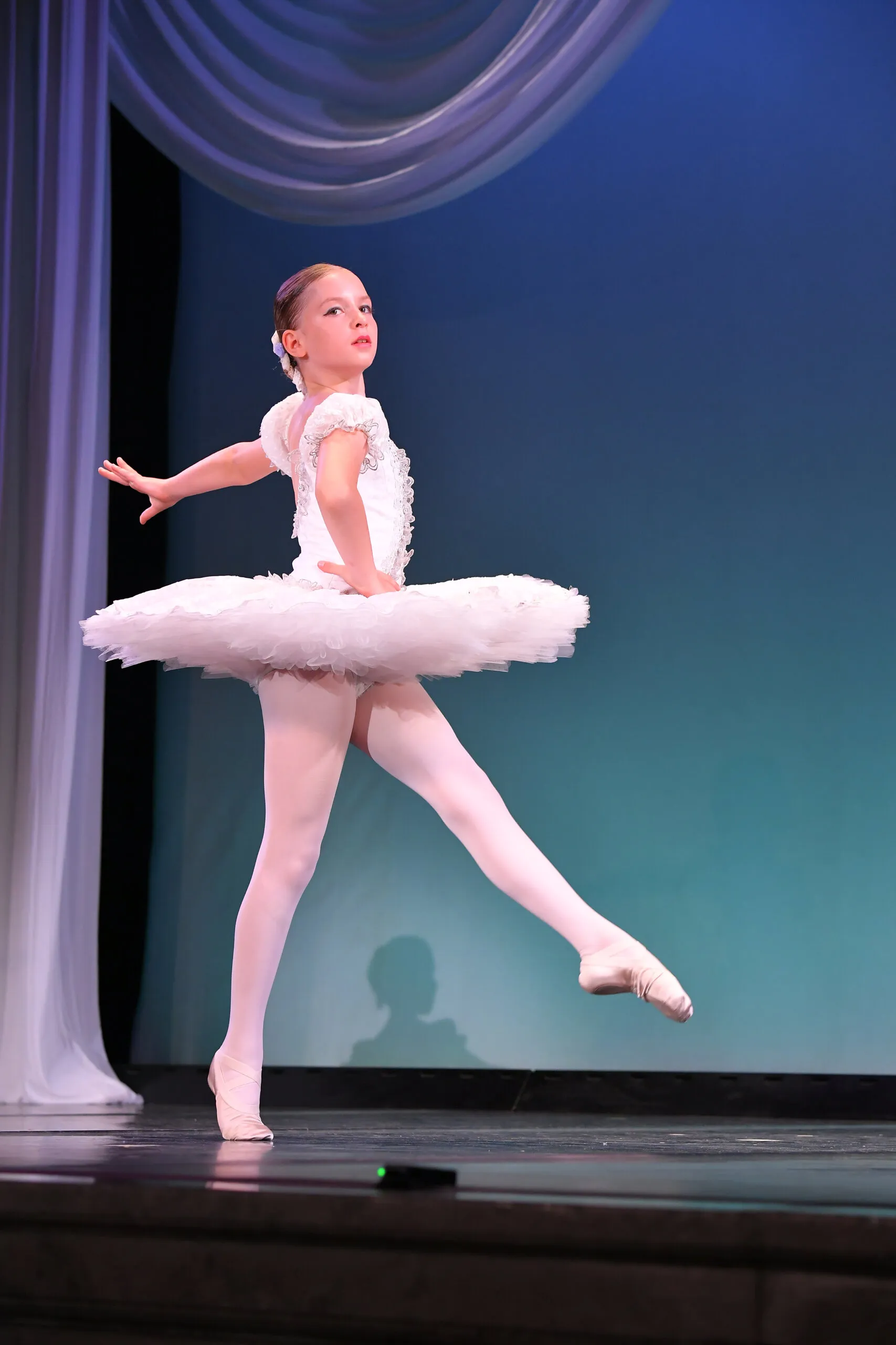 Une jeune élève en tutu blanc, figure entière sur le fond bleu.