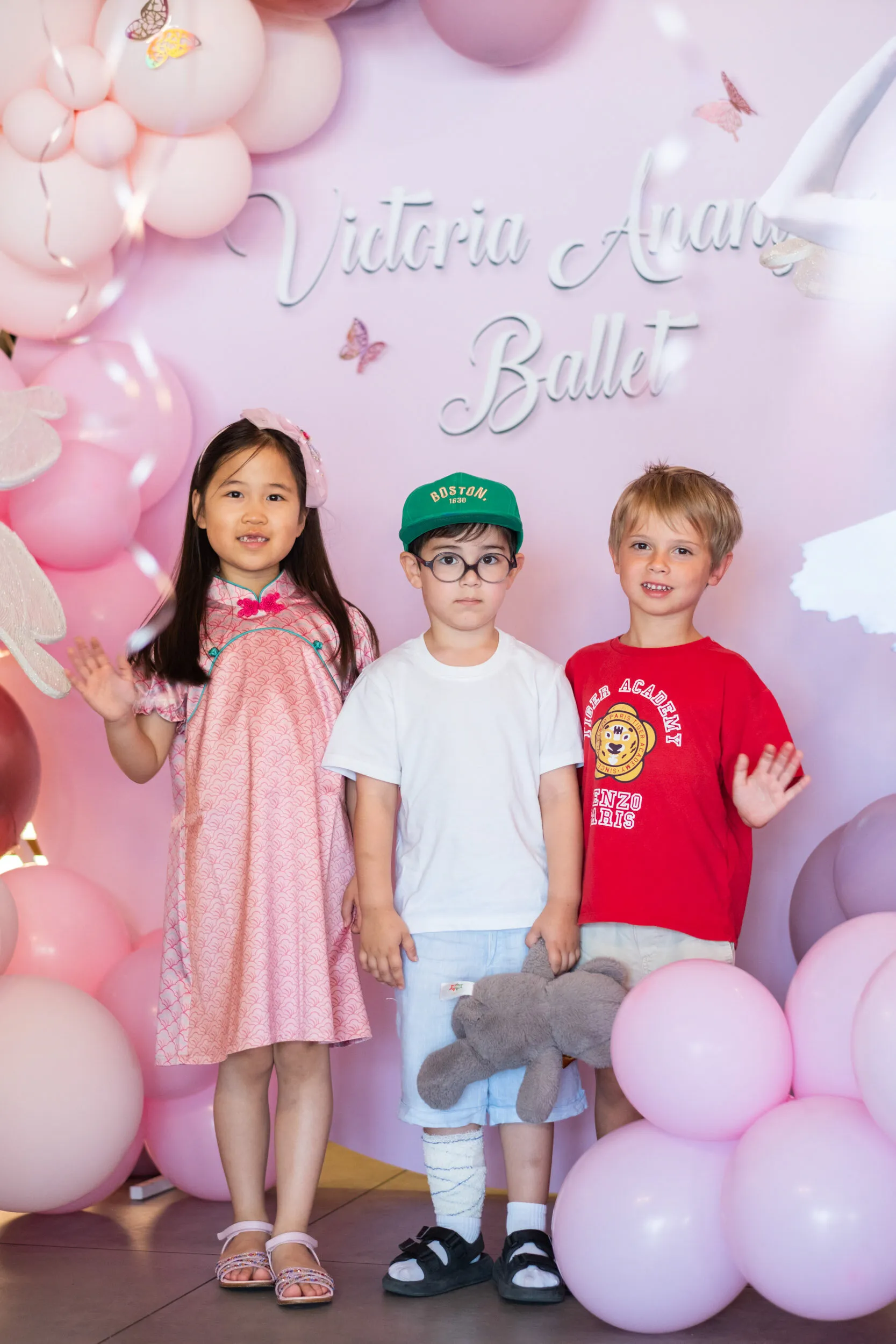 Three children in front of the party backdrop, under an arch of pink balloons.