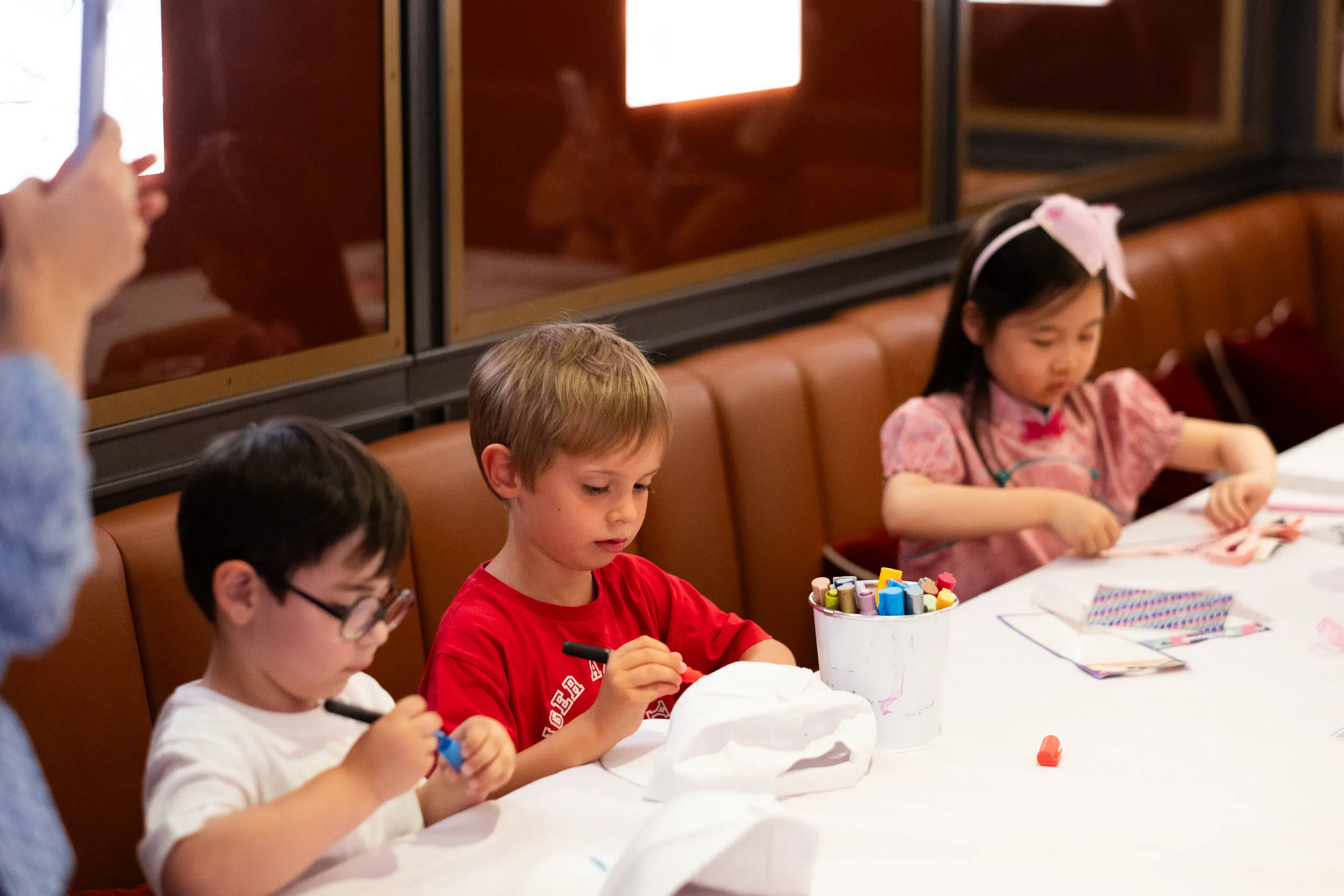 Three children concentrate on decorating their caps at the workshop table.
