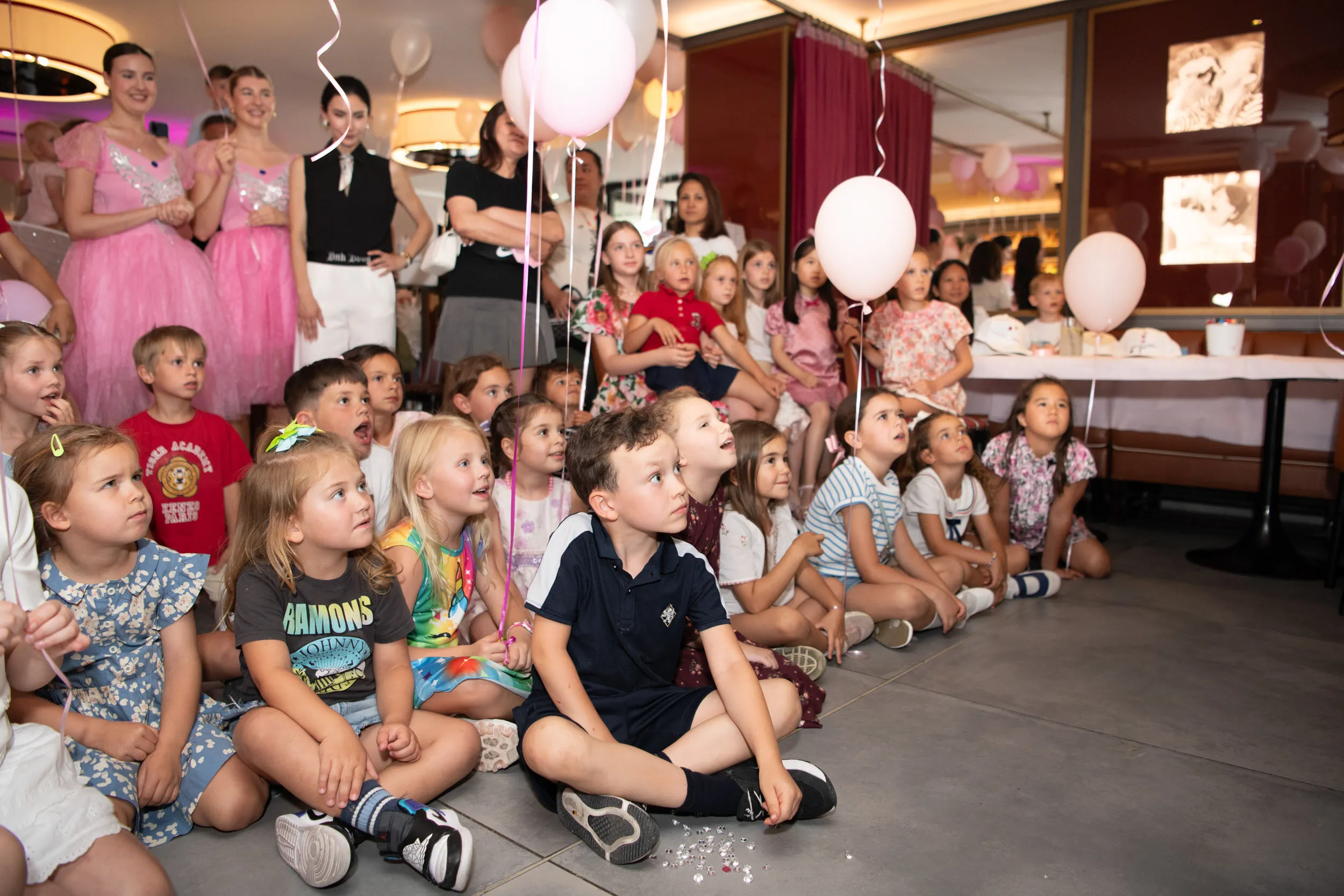 The children sit cross legged watching the show, with families around them.