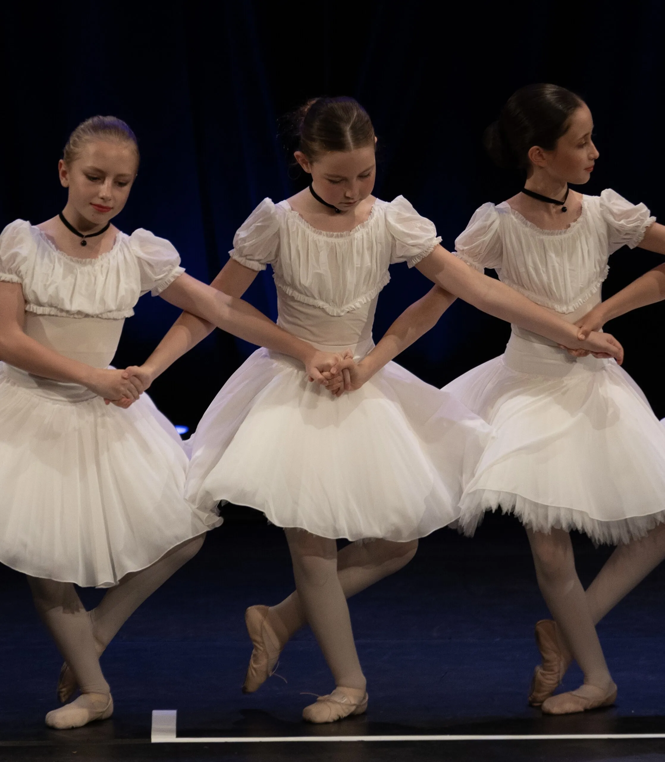 Trois élèves en tutus romantiques blancs, mains réunies, sur le plateau bleu.