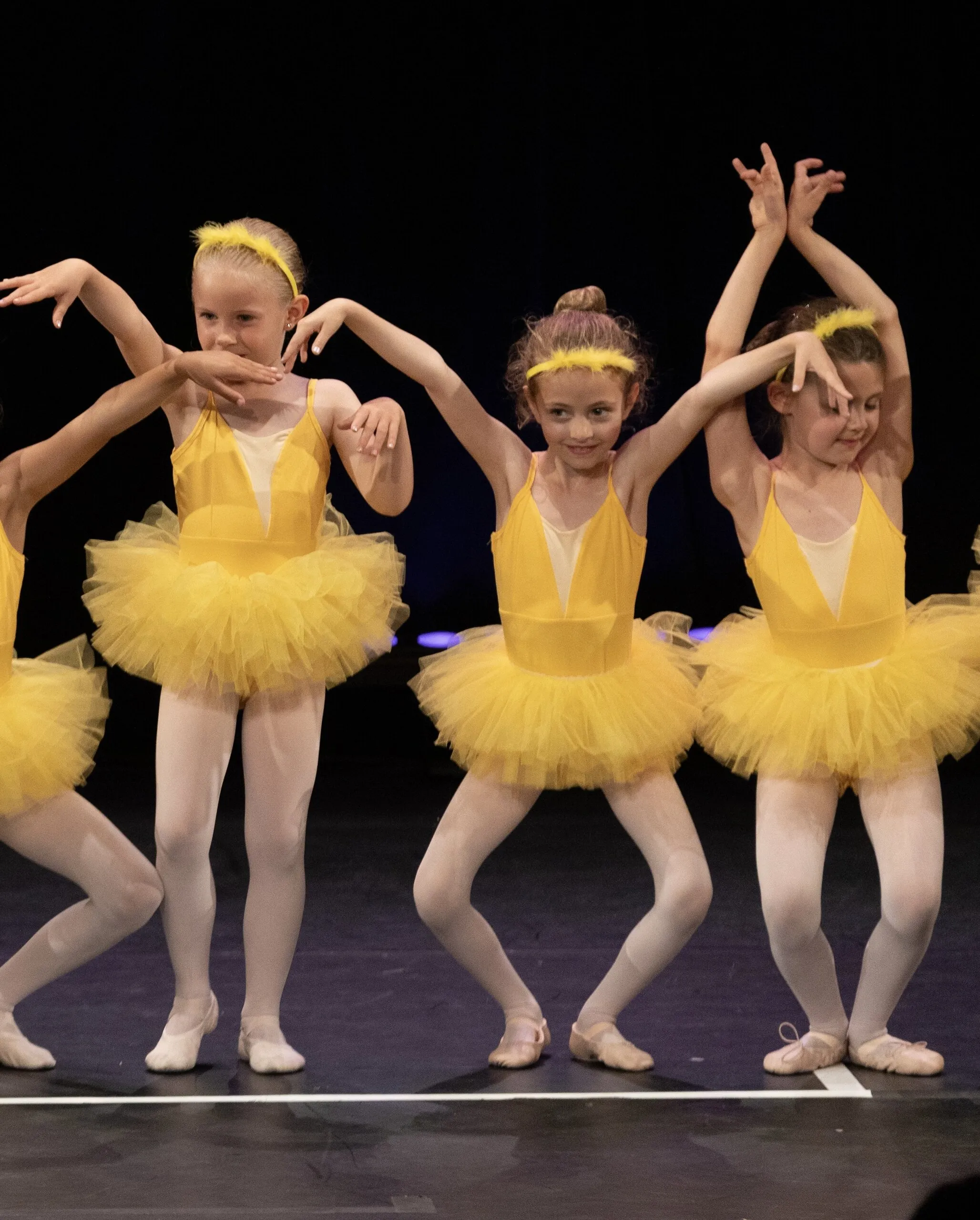 Trois petites en tutus jaunes, les bras arrondis au-dessus de la tête.