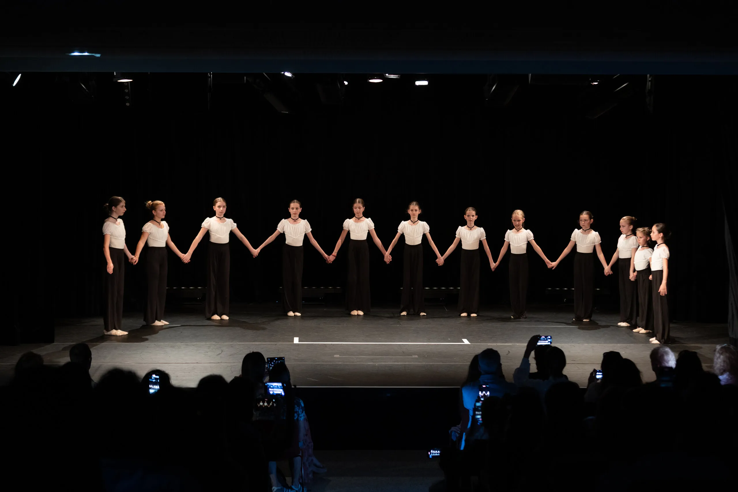 Une ligne de danseurs en hauts blancs et pantalons sombres, main dans la main.