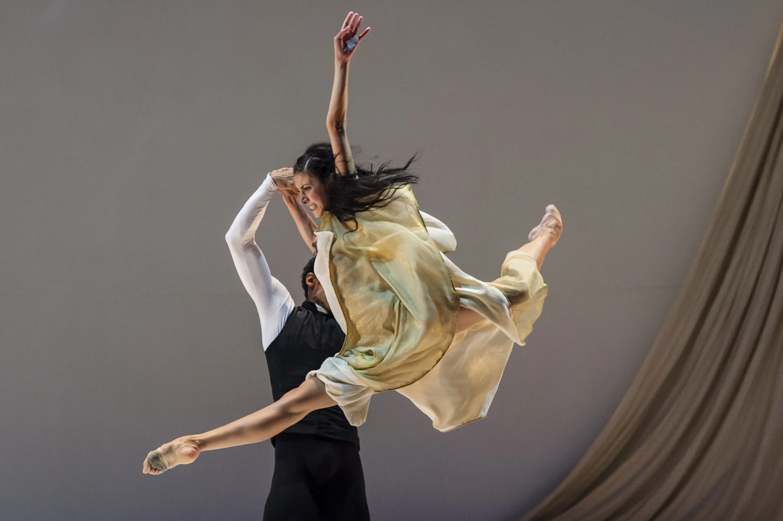 Victoria portée en plein saut par son partenaire, la robe dorée en mouvement.
