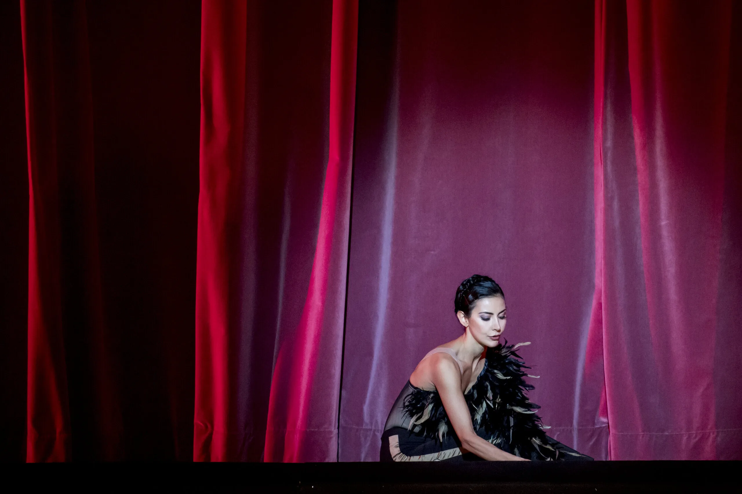 Victoria assise dans la lumière d'un rideau rouge, en costume de plumes noires.