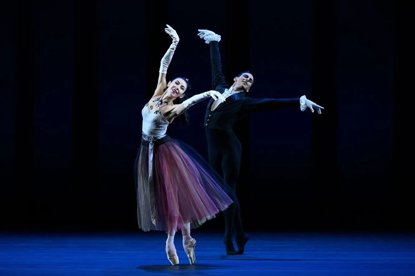 Victoria sur pointes en jupe de tulle mauve, le bras levé, aux côtés de son partenaire sur une scène bleue.