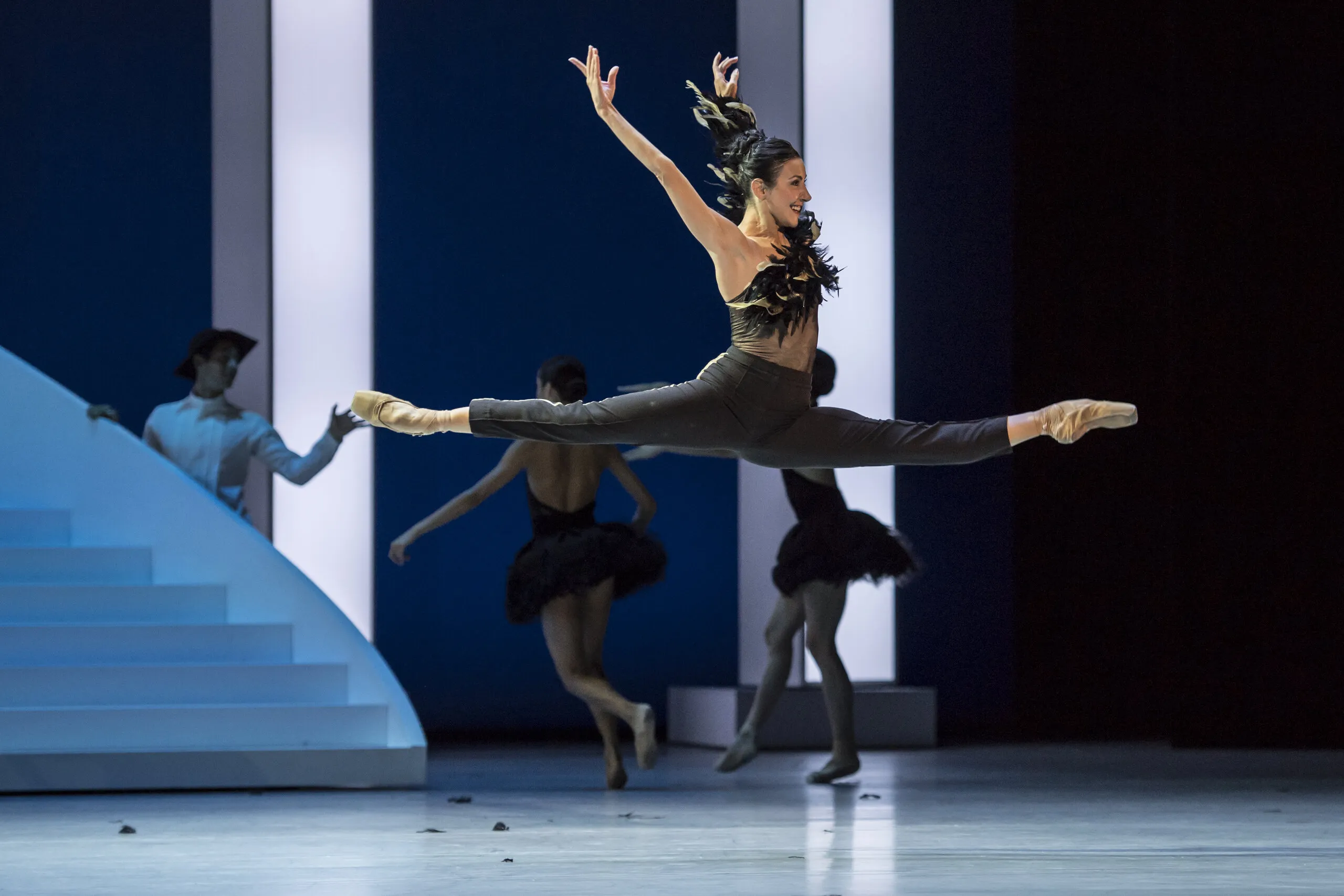 Victoria en grand jeté devant l'escalier blanc du décor, deux danseuses en arrière-plan.