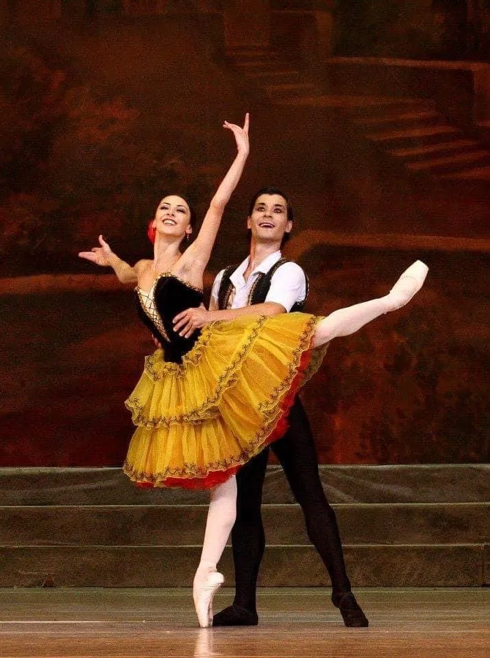 Victoria en tutu jaune et rouge, sur pointes, la main dans celle de son partenaire.