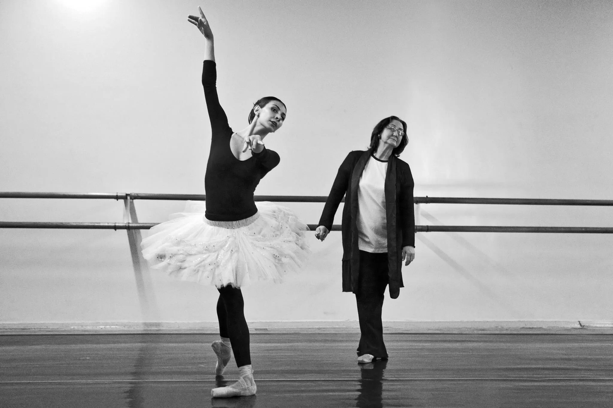 Victoria en tutu blanc à la barre, le bras levé, aux côtés d'une femme en tenue de ville, en noir et blanc.