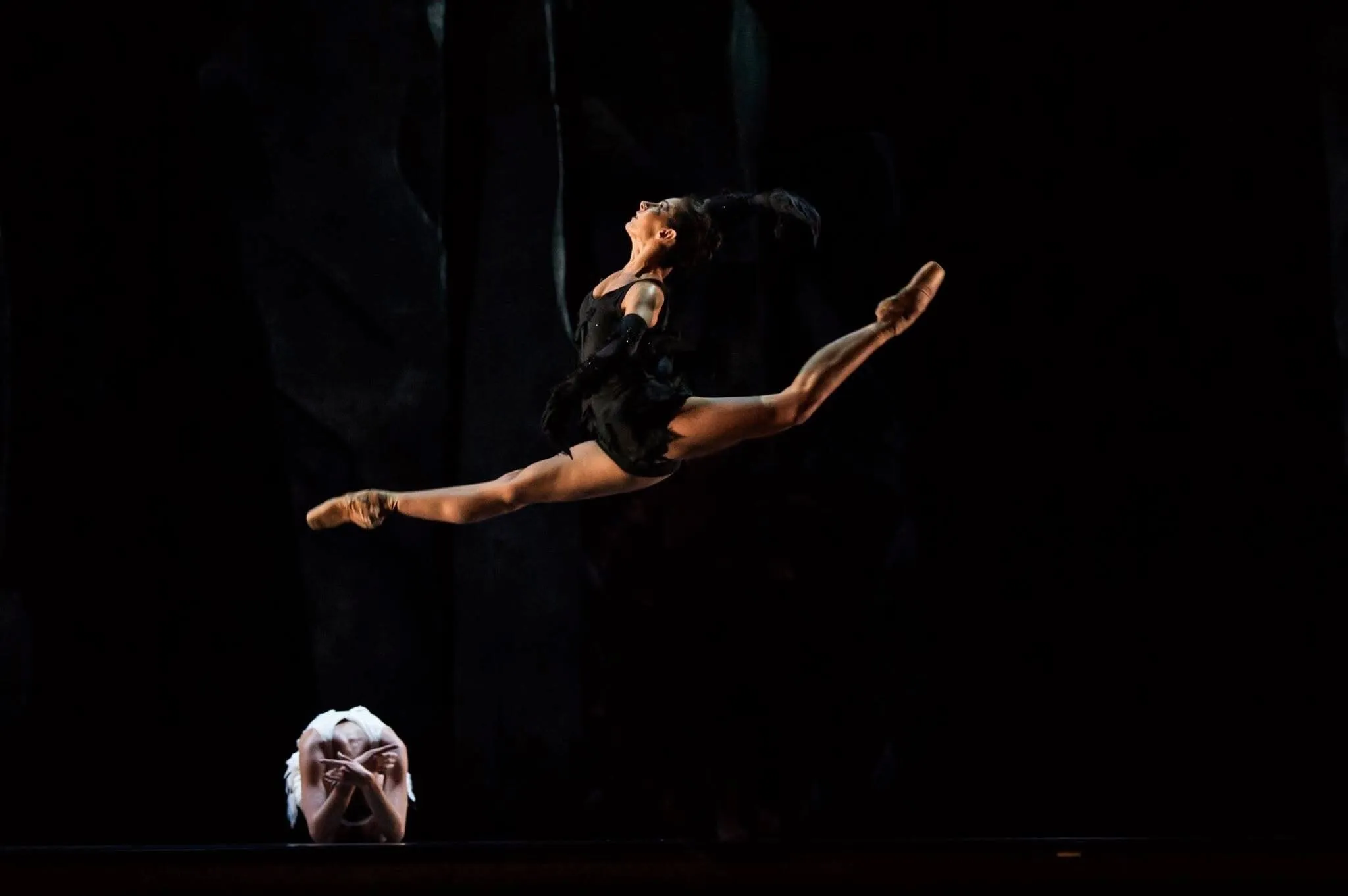 Une danseuse en plein grand jeté sur une scène sombre, un masque blanc posé au sol.