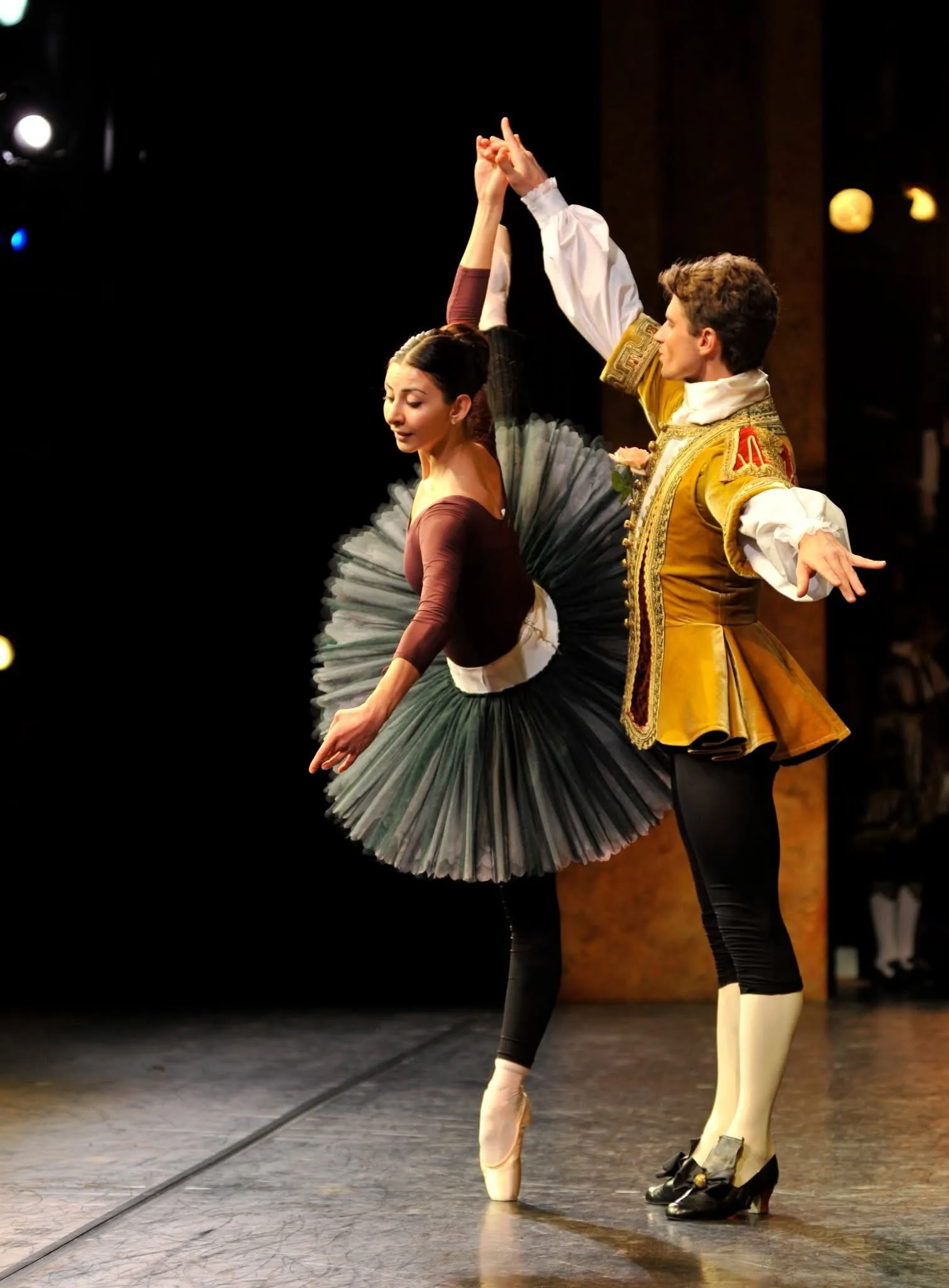 Victoria en tutu gris et corsage bordeaux, la main dans celle de son partenaire en habit doré.