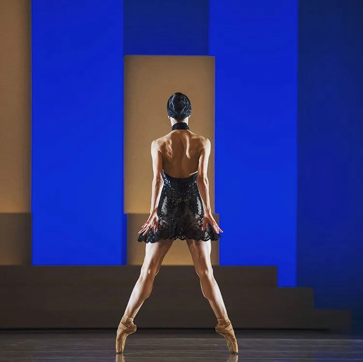 Une danseuse de dos sur pointes, en corsage noir brodé, devant un décor bleu à degrés.