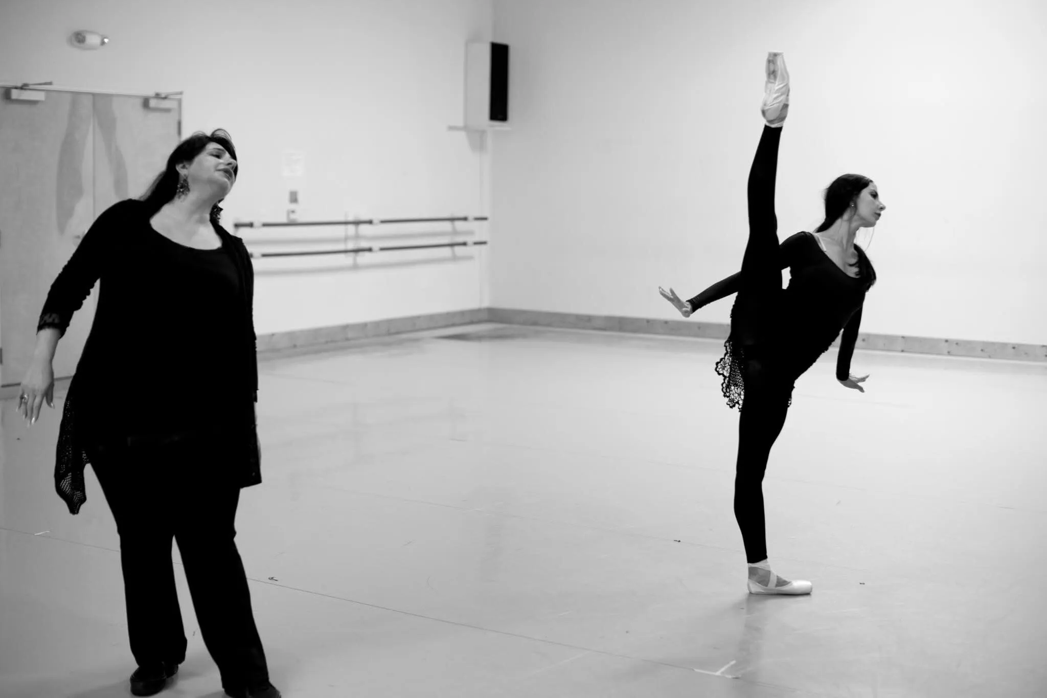 Dans un studio clair, une répétitrice suit une danseuse en arabesque, en noir et blanc.